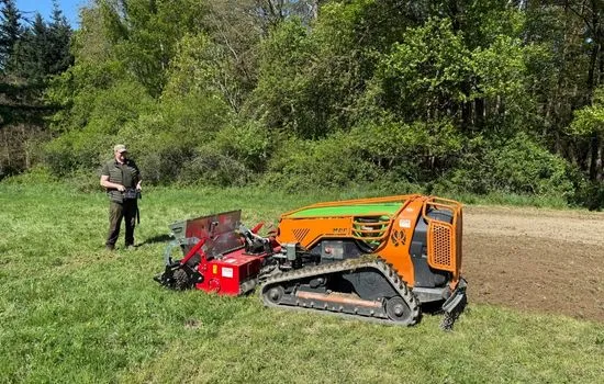 Funkraupe mit rotem Anbaugerät auf einer Wiese, Bediener daneben